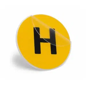 Pegatina circular amarilla con la letra H mayúscula en negro para vehículos históricos, mostrando el plástico transparente transportador medio despegado.