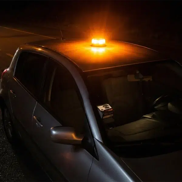 Baliza V16 conectada Metalight emitiendo una potente luz ámbar intermitente sobre el techo de un coche aparcado en la carretera de noche.